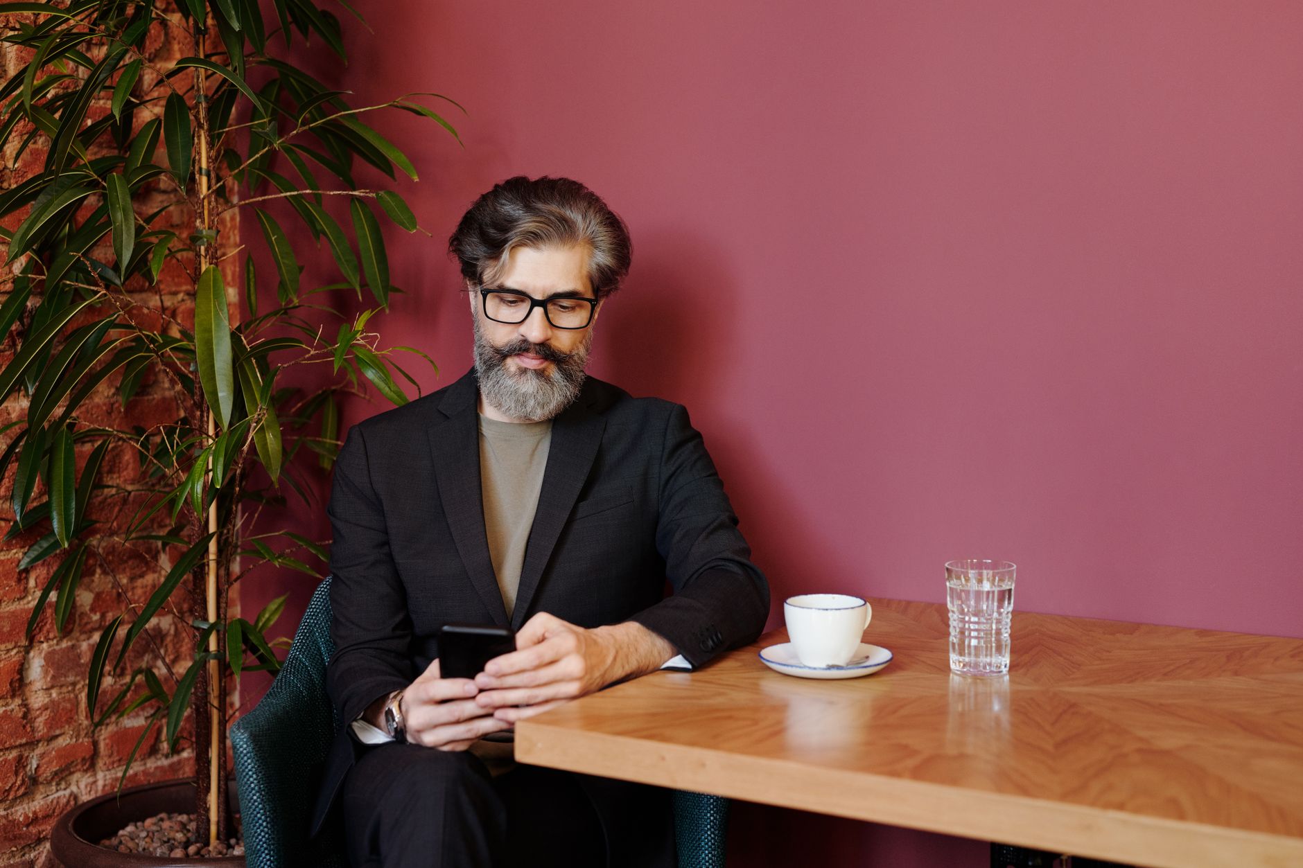 Homme à lunettes et à la barbe grisonnante, assis à une table de café, qui consulte son téléphone devant une tasse et un verre d’eau