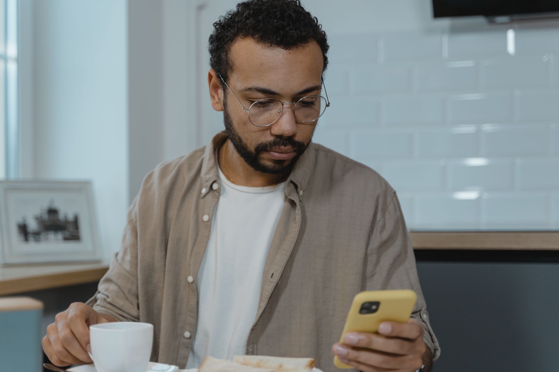 Homme à lunettes rondes qui lit une invitation sur son téléphone jaune en prenant son petit-déjeuner