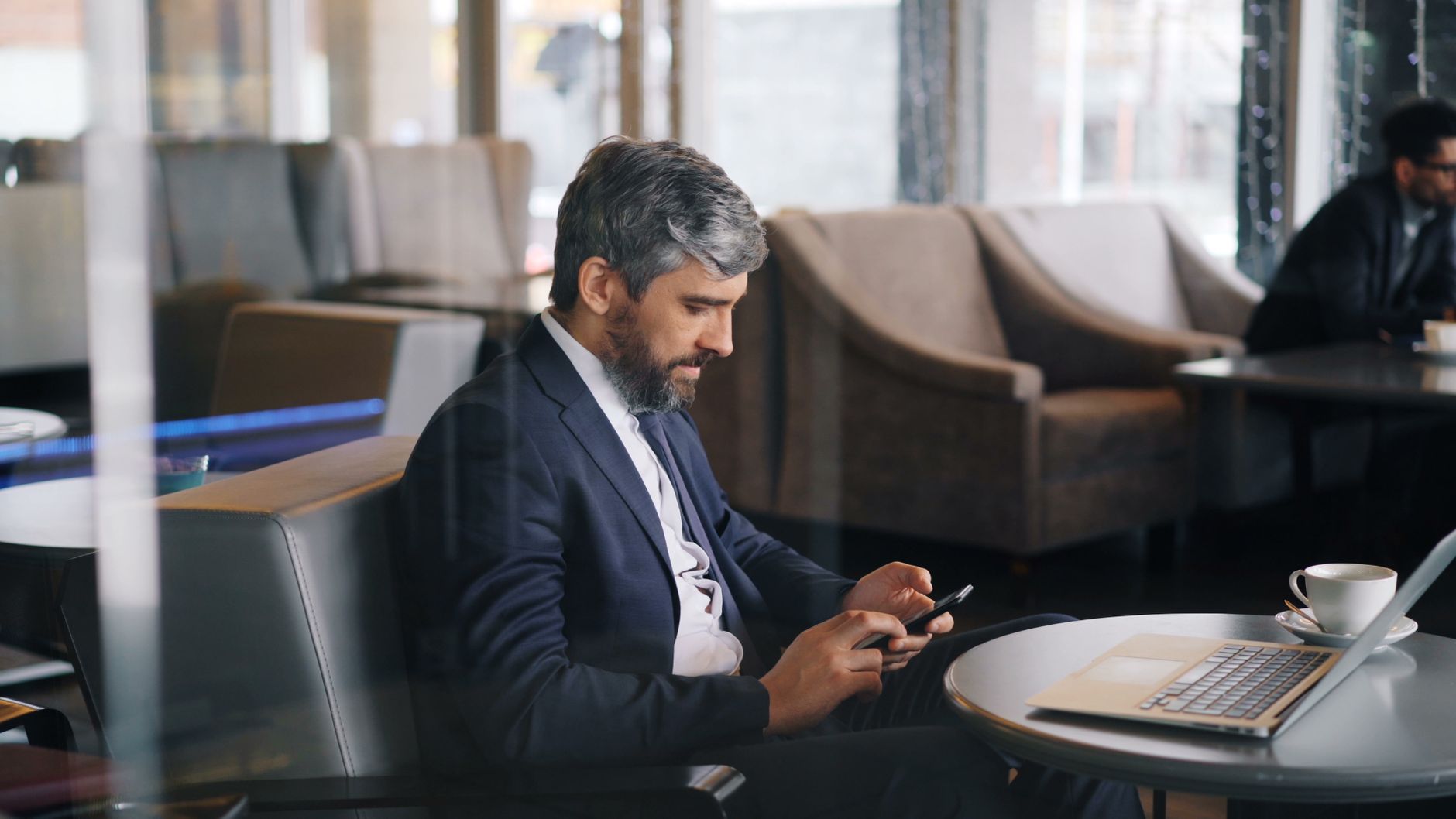 Homme en costume assis dans un café, qui répond sur son téléphone, un ordinateur portable et une tasse posés sur la table