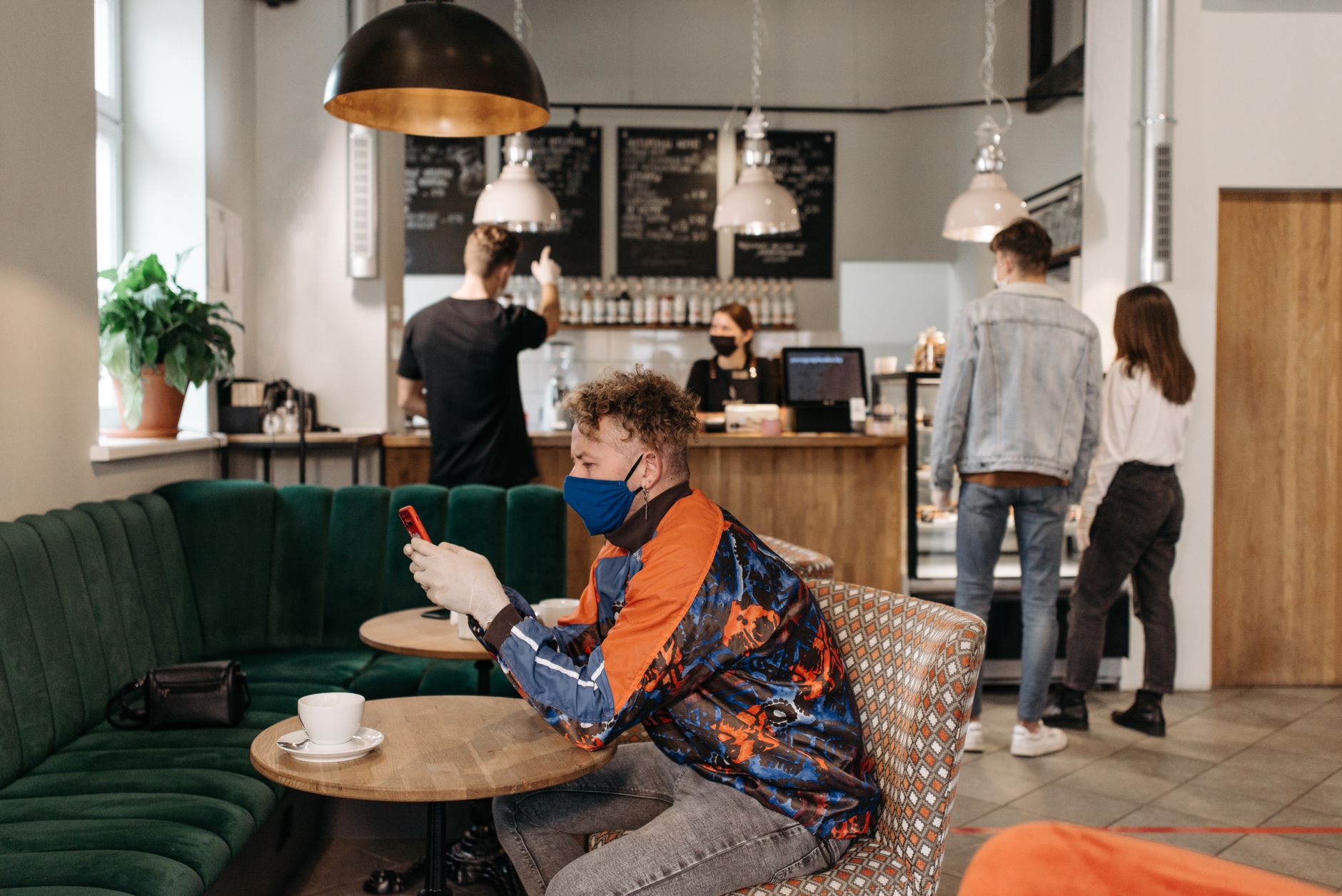 Un client assis dans un café, masque sur le visage, qui regarde son téléphone pendant que d’autres personnes patientent au comptoir
