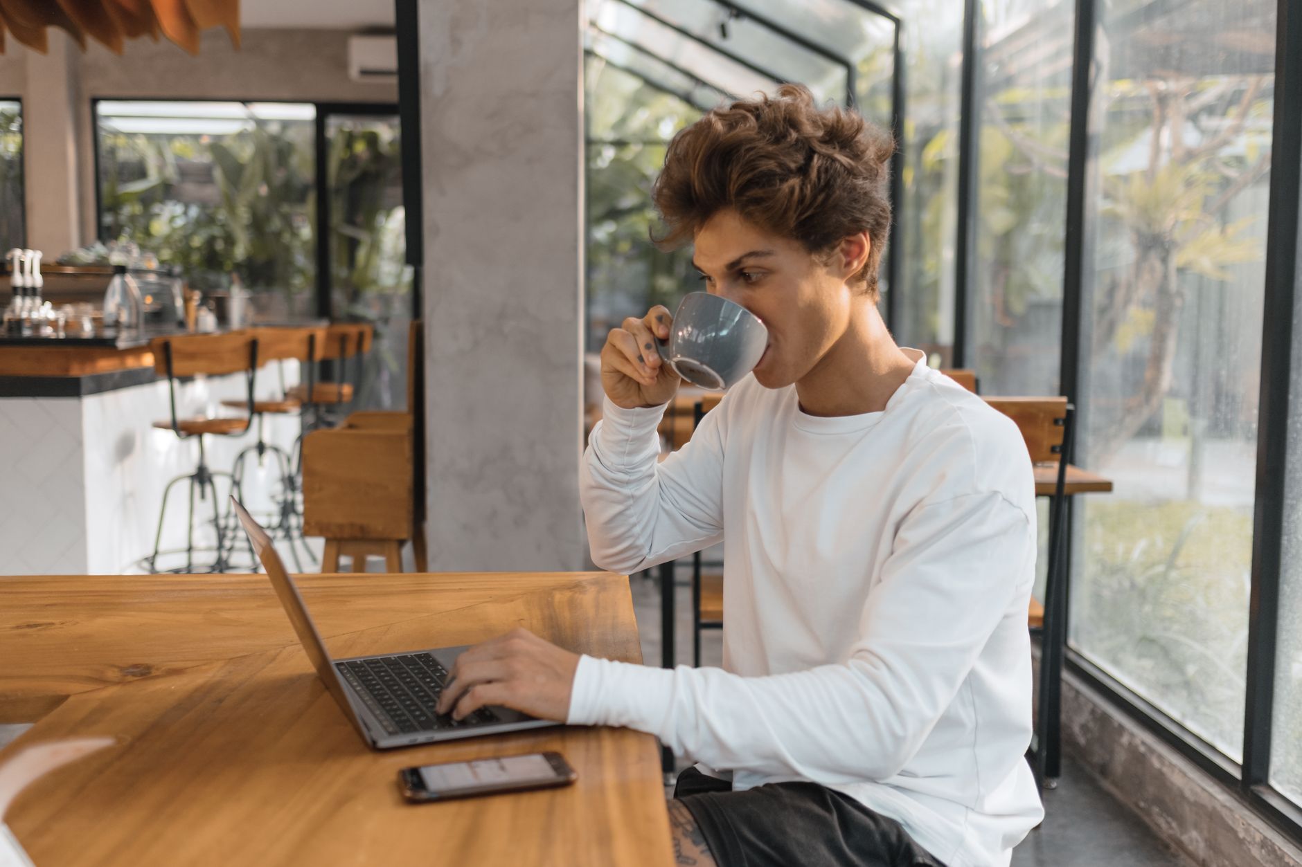 Jeune homme qui boit un café devant son ordinateur portable, à une grande table en bois dans un café lumineux